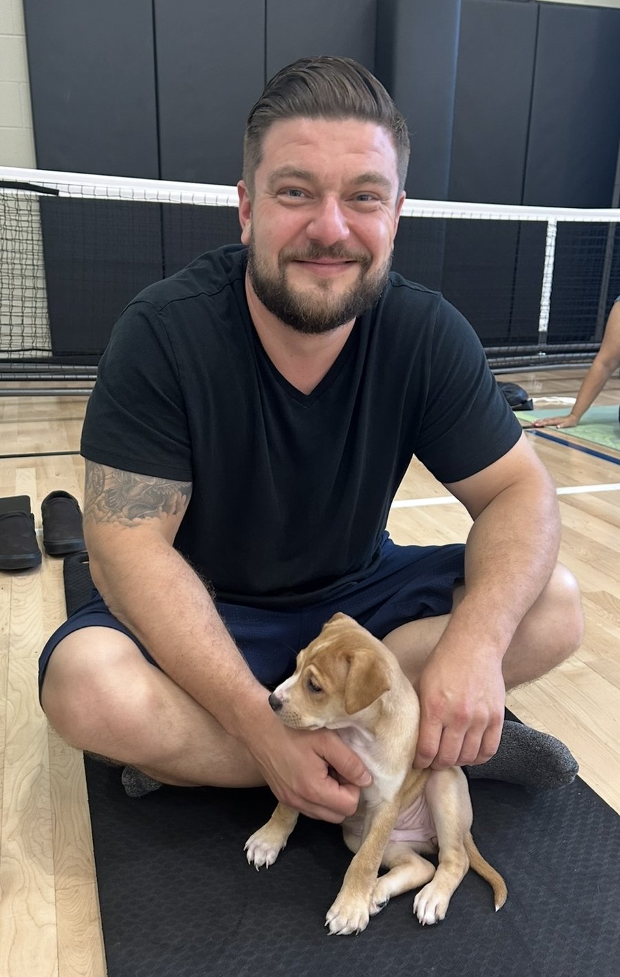 Markian sitting cross-legged on a gym floor, smiling, with his puppy Twinkle in his lap